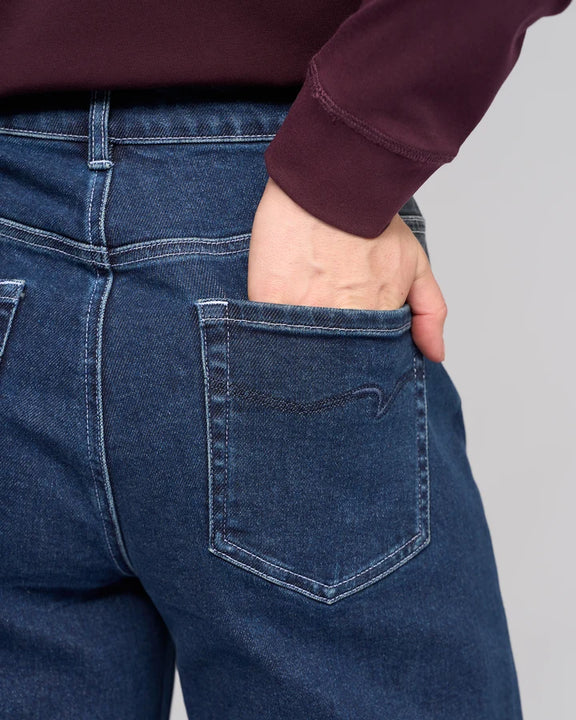 Close-up back view of a person wearing dark blue denim jeans, with one hand tucked into the back pocket. Vassalli Alley Jean in dark blue denim.
