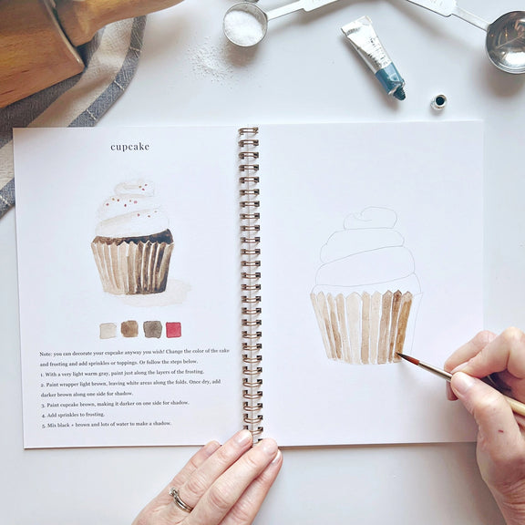 Open book with cupcake illustration and hands using a paintbrush