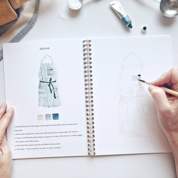 Person holding a paintbrush over a book with apron design illustrations on a white surface.