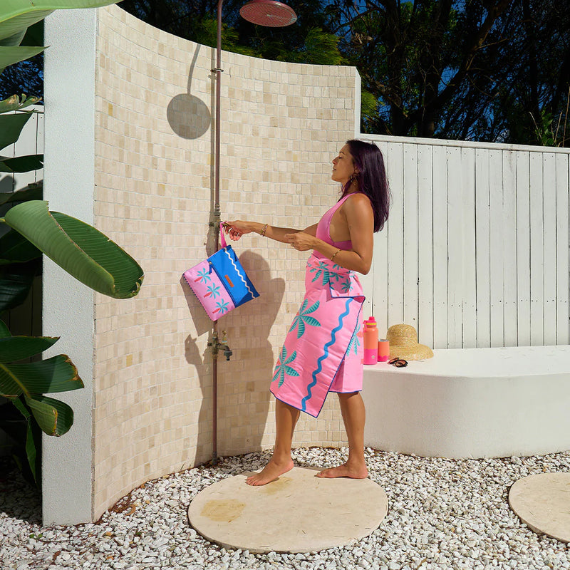 Woman in a pink dress with blue patterns standing under an outdoor shower holding colorful bags.