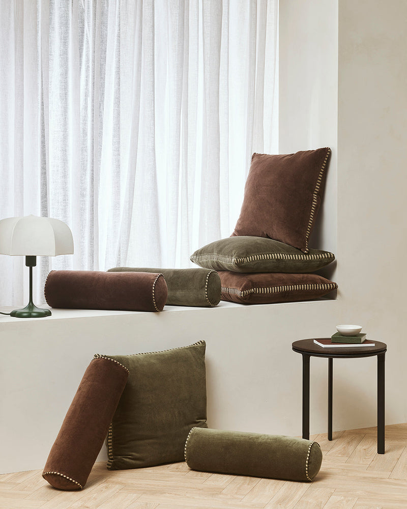 Brown and green cushions on a white surface with a lamp and small table in the background.