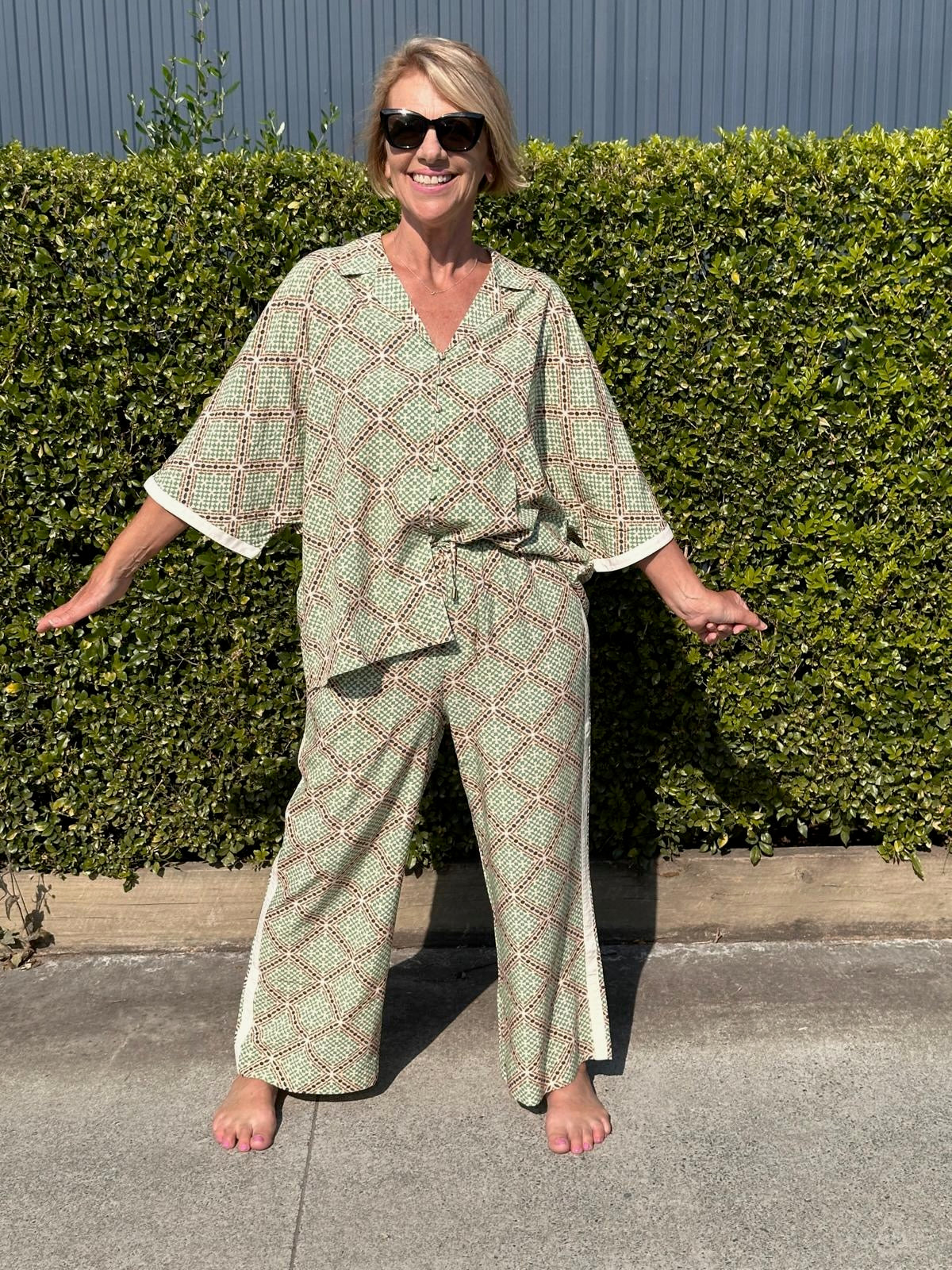 Woman wearing a green patterned outfit standing in front of a hedge.