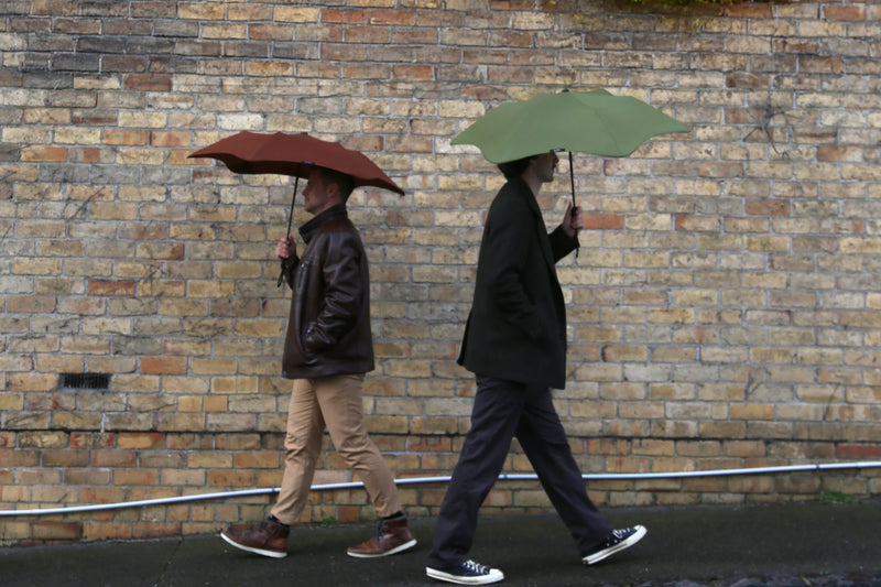 Two people walking side by side with umbrellas against a brick wall.