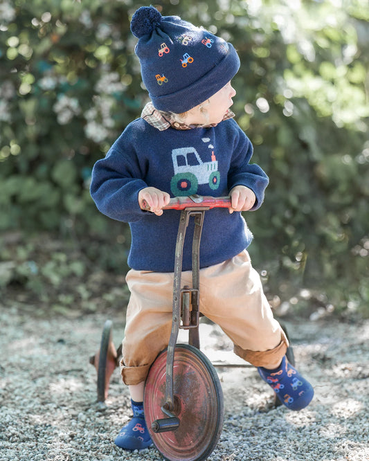 Child wearing a blue sweater with a tractor design, beige pants, and blue boots, standing on a gravel path with greenery in the background.