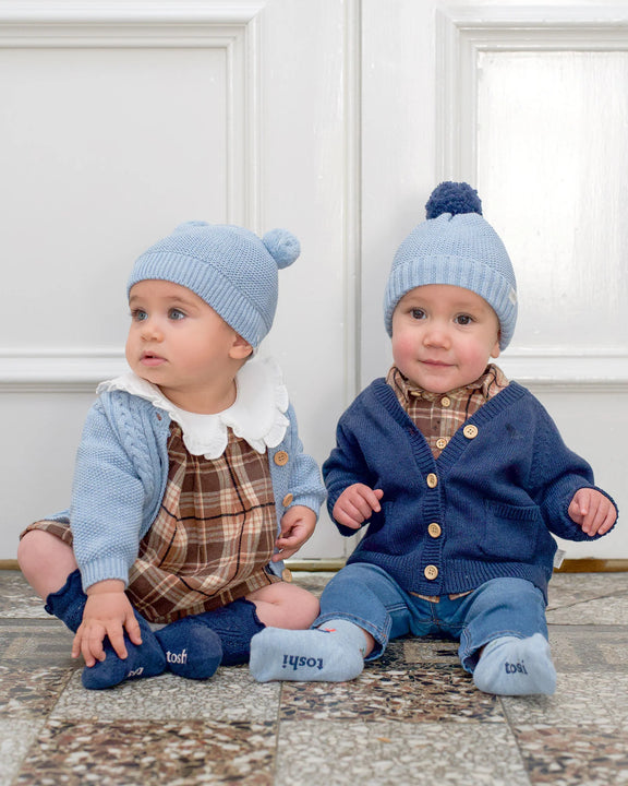Two babies wearing blue knit hats and plaid outfits sitting on a rug. Baby boy wearing Toshi Organic Cardigan Remy Ink