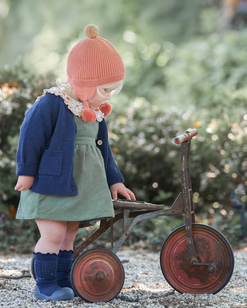 Child in a green dress and blue sweater with a pink hat playing with an old tricycle outdoors. Sweater is a Toshi Organic Cardigan Remy Ink