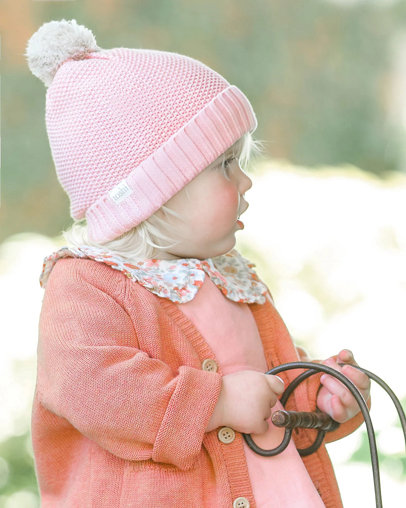 Child wearing a pink knit hat with a pom-pom and an orange cardigan outdoors. Toshi Organic Cardigan Remy Tumbleweed