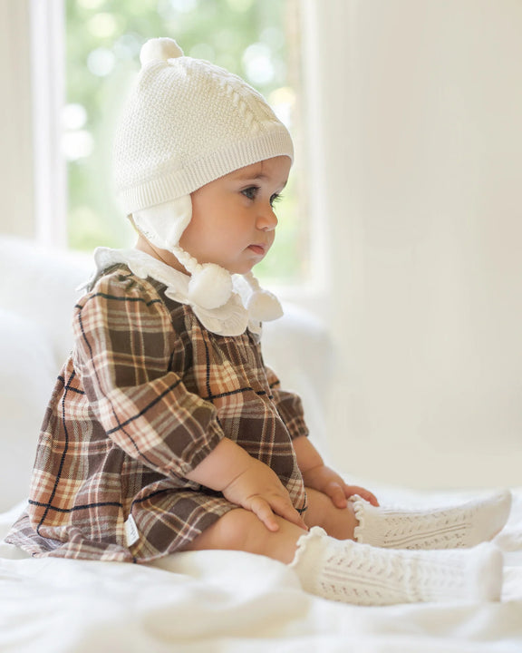 Baby wearing a plaid dress, white knit hat, and matching socks sitting on a white surface.