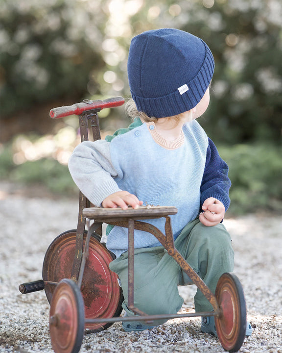 Child sitting on a vintage tricycle outdoors with a blurred natural background wearing Blue organic cotton baby pullover