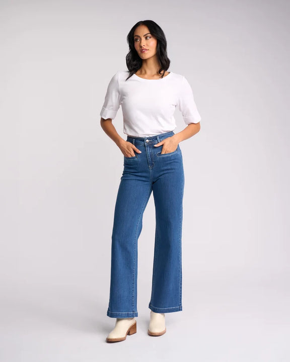 A woman modeling a pair of high-waisted, wide-leg blue jeans with a white t-shirt and white heeled boots, standing against a light gray studio background.