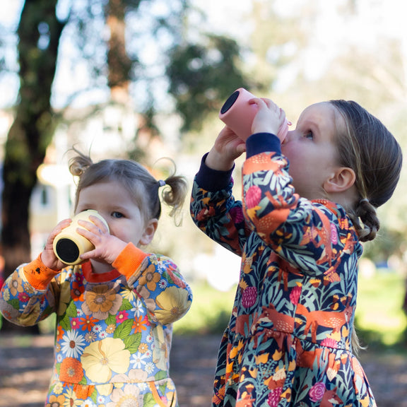 Two children in colorful outfits drinking from Made By Fressko piccolo cups outdoors.
