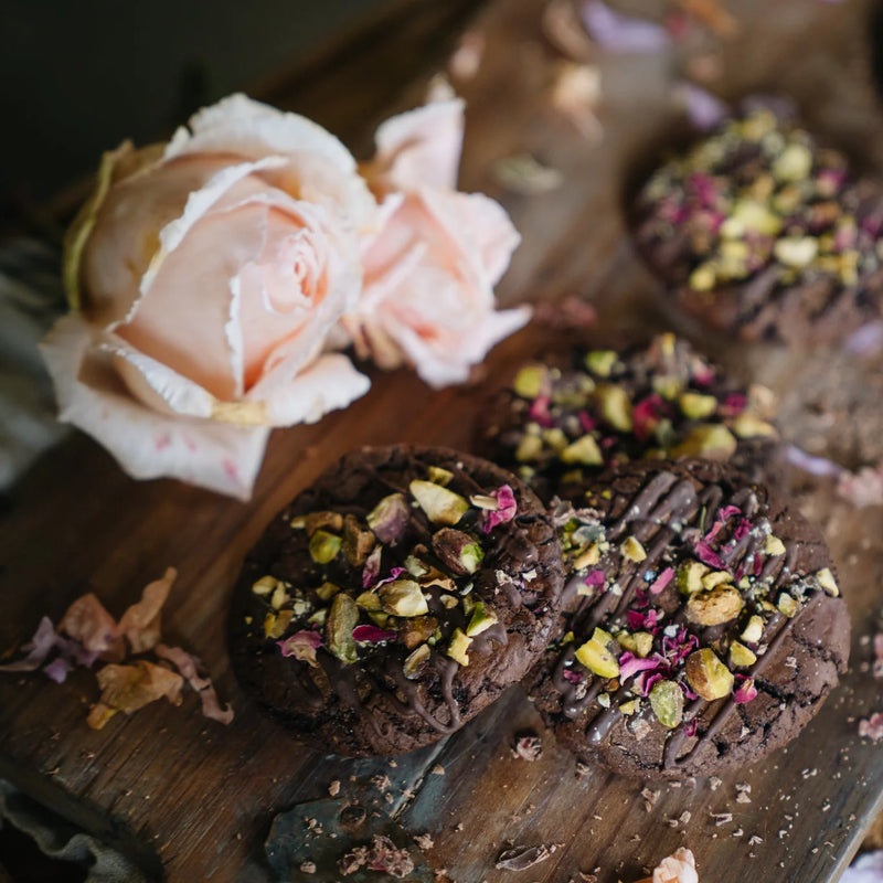 Chocolate cookies with pistachio and dried flower toppings on a wooden board with a pink rose.