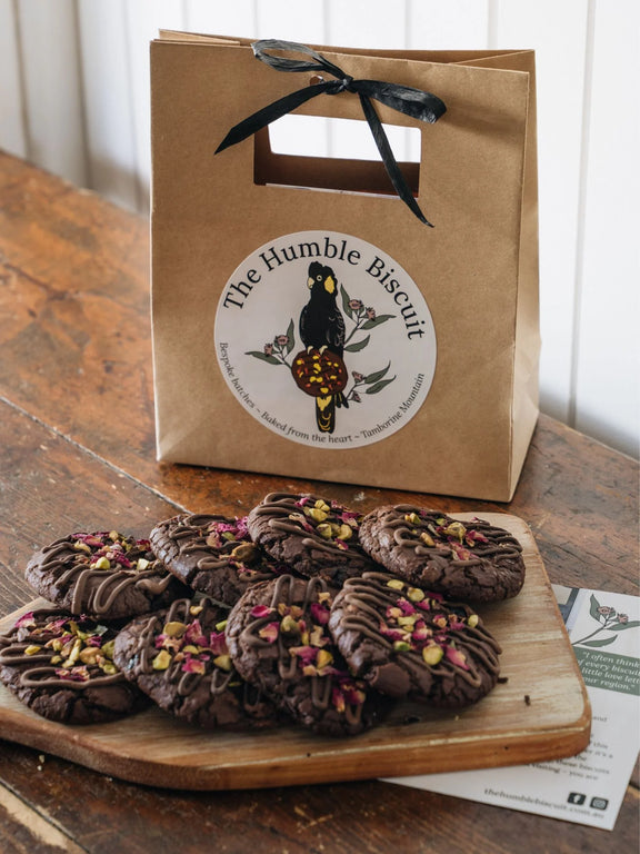 Chocolate cookies with dried flowers on a wooden board next to a 'The Humble Biscuit' paper bag.