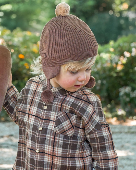 Child wearing a brown knitted hat with earflaps and plaid coat outdoors.