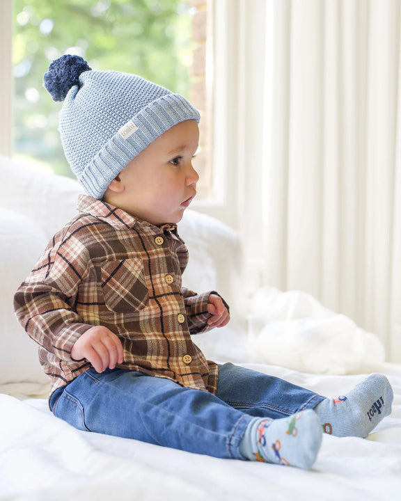 Baby wearing a blue knit hat with a pom-pom, plaid shirt, and jeans sitting on a white surface.