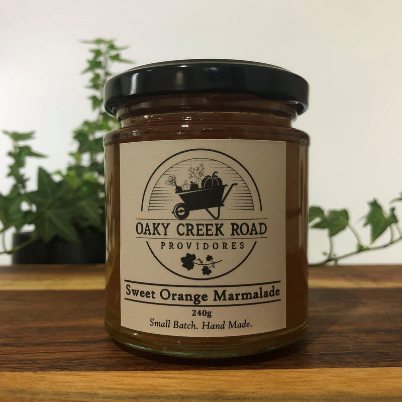 A jar of Sweet Orange Marmalade with a label that reads 'OAK CREEK ROAD PROVIDORES' and 'Sweet Orange Marmalade 240g'. The jar is placed on a wooden surface with a plant in the background.