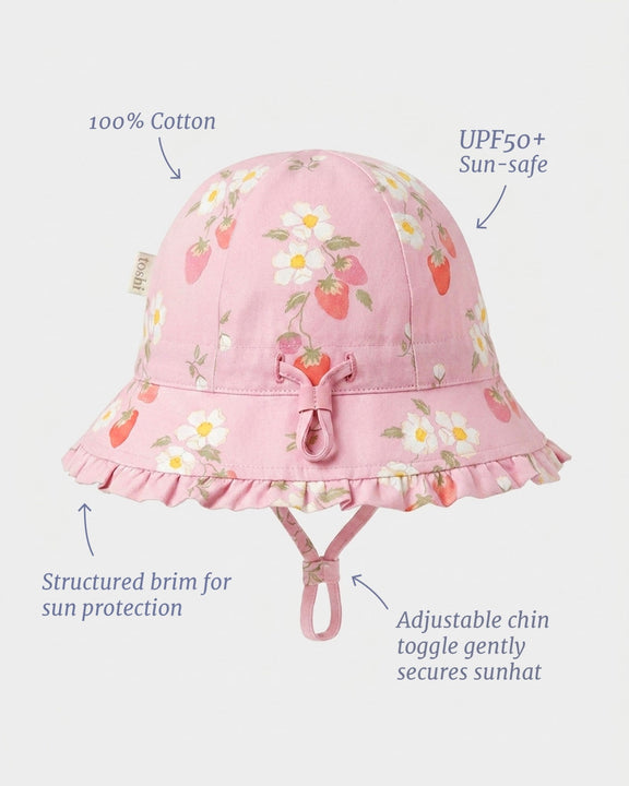 Pink cotton sun hat with a strawberry and daisy floral print, featuring a ruffled brim, chin tie, and text callouts highlighting fabric and sun protection features.