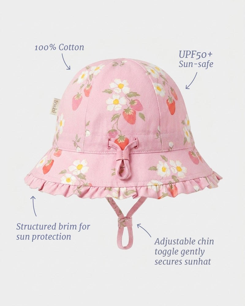 Pink cotton sun hat with a strawberry and daisy floral print, featuring a ruffled brim, chin tie, and text callouts highlighting fabric and sun protection features.