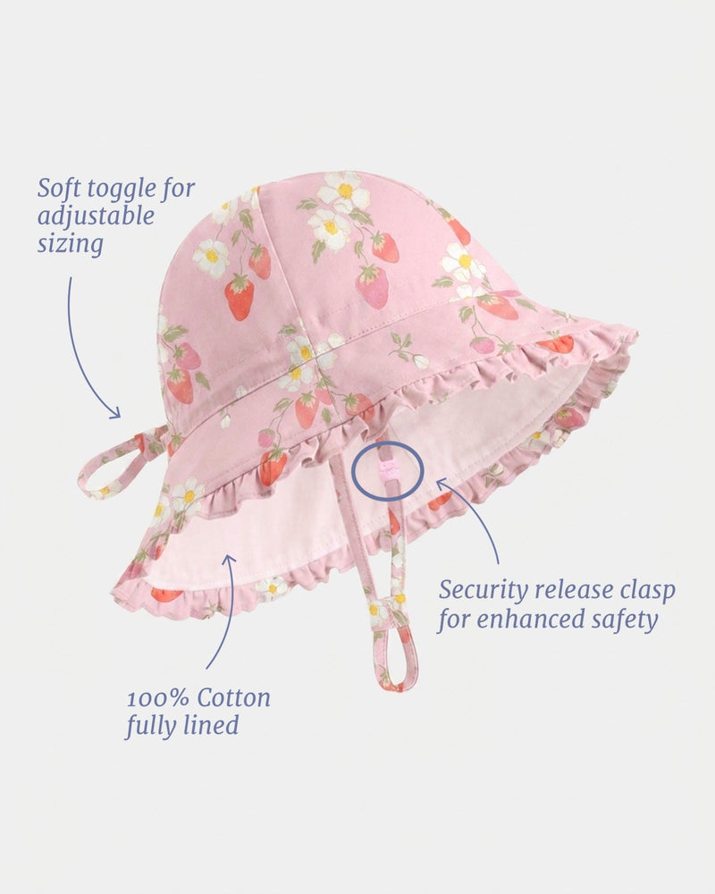 Pink cotton sun hat with a strawberry and daisy floral print, featuring a ruffled brim, adjustable tie, and safety release clasp, shown with callout text and arrows on a light gray background.