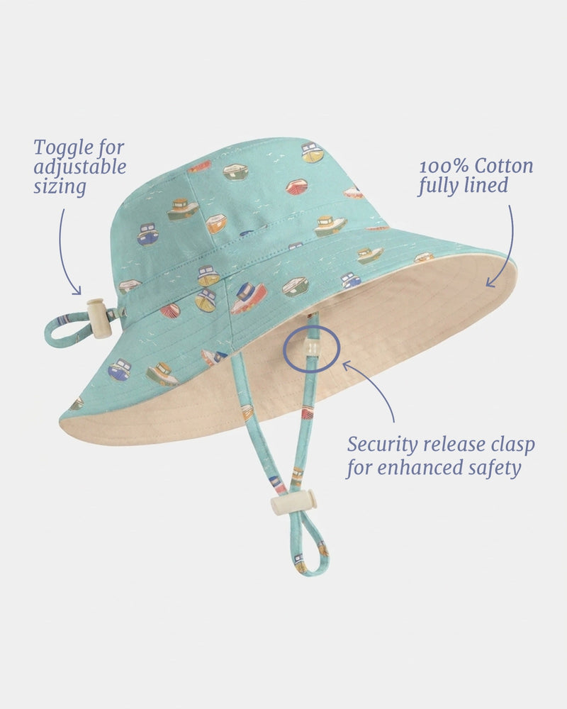 A light blue cotton sun hat with a colorful beach-themed print, featuring text callouts highlighting adjustable sizing, fully lined interior, and a safety release clasp, set against a solid light gray background. Australian designed Toshi Tide Tugboat sun hat with broad protective brim.