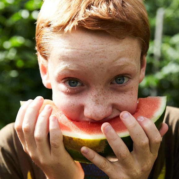 Child eating a watermelon slice outdoors with greenery in the background