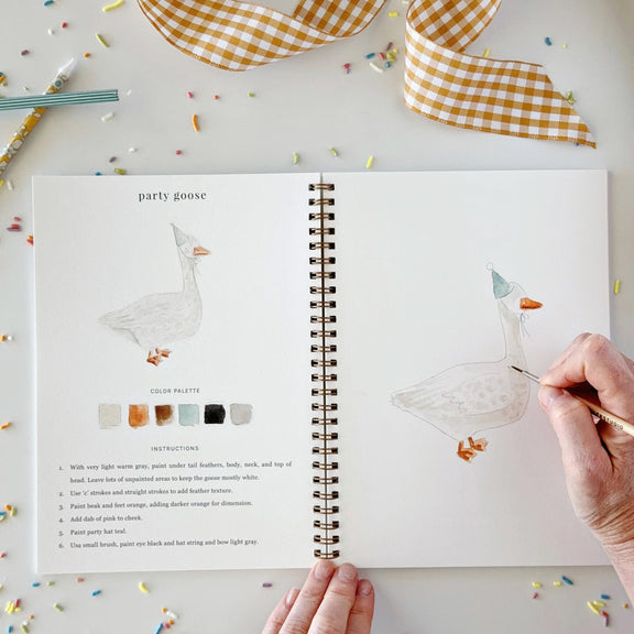 An open spiral-bound art journal or coloring book on a white surface, showing illustrated goose designs, color palettes, and instructions. A hand is coloring one goose with a gold pen, and another hand holds the bottom edge of the page.