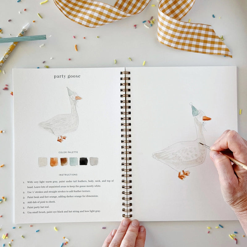 An open spiral-bound art journal or coloring book on a white surface, showing illustrated goose designs, color palettes, and instructions. A hand is coloring one goose with a gold pen, and another hand holds the bottom edge of the page.