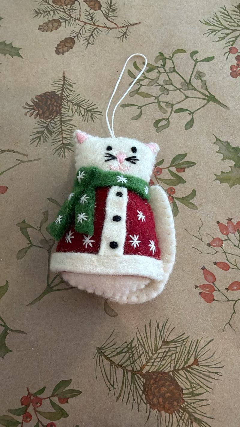 A handmade felt Christmas ornament shaped like a cat wearing a red sweater and green scarf, resting on a brown kraft paper background with a pine and berry pattern.